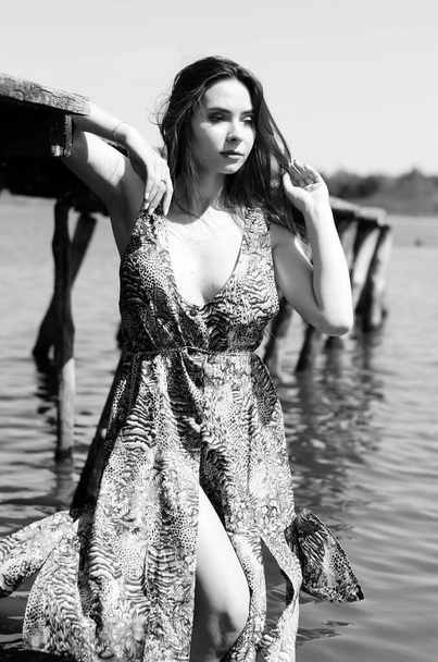 Ciemnowłosa kobieta pozująca na drewnianym pomoście nad jeziorem. Kobieta w letnim stroju z plecami odwróciła się do drewnianej kładki nad jeziorem Neusiedl. Odbicia słońca w hangarze na łodzi w tle - Zdjęcie, obraz