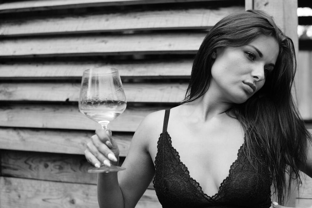 Pretty young brunette woman dressed in dark lingerie holding glass of wine and posing on leather sofa near wooden house outdoor  - Foto, Imagem