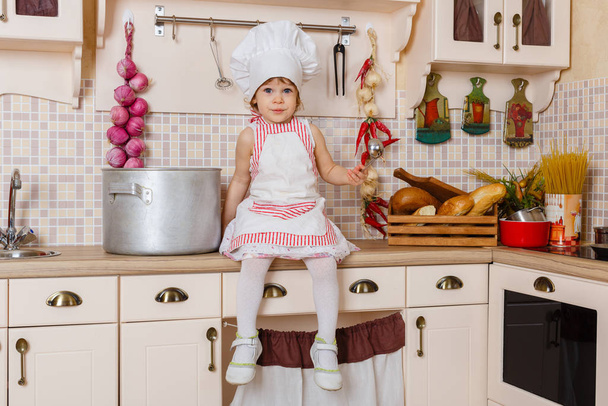 Little girl in apron and cap of the cook sitting  in the kitchen in the house. Mother's helper. 2 year old. - 写真・画像