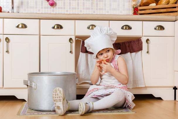 Little girl in apron and cap of the cook sitting  in the kitchen in the house. Mother's helper. 2 year old. - Fotoğraf, Görsel