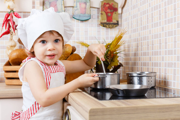 Little girl in apron and cap of the cook stands in the kitchen near cooker in the house. Mother's helper. 2 year old. - Fotoğraf, Görsel