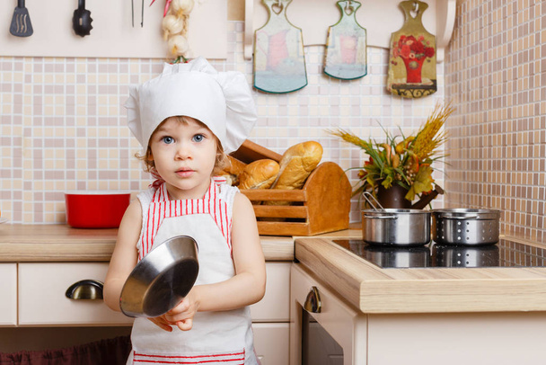 Little girl in apron and cap of the cook stands in the kitchen near cooker in the house. Mother's helper. 2 year old. - Foto, imagen