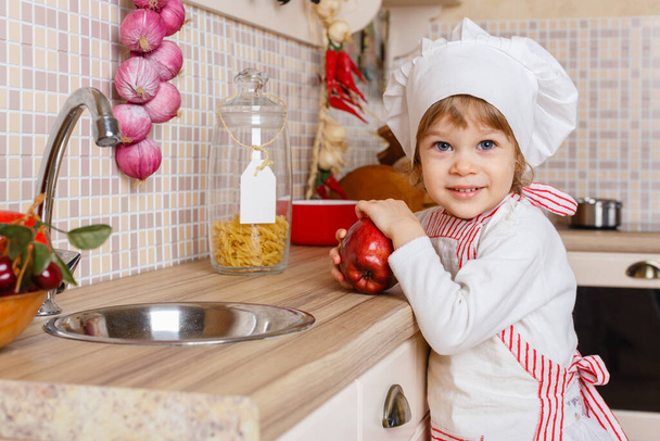 Little girl in apron and cap of the cook with an apple stands in the kitchen near sink at home. Mother's helper. 2 year old. - Foto, Bild