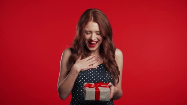 Joyful beautiful woman with perfect makeup holding gift box with bow on red wall background. Retro styled girl smiling, she is glad to get present. - Кадри, відео