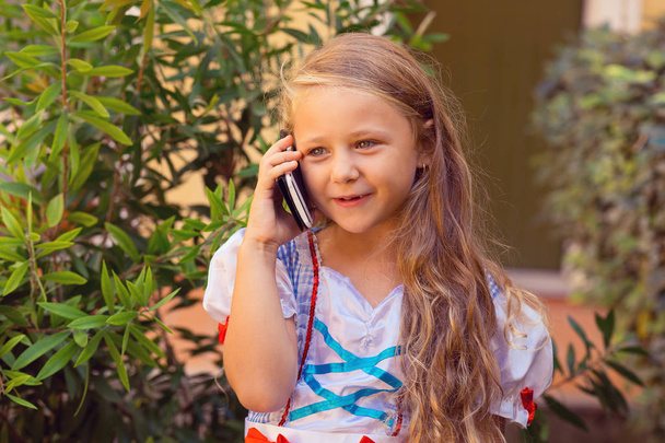 Phone talk happy. Little girl talking on the phone, excited and happy explaining something to her mother nanny father parents isolated green shrubs nature near house outdoors on the background - Photo, Image