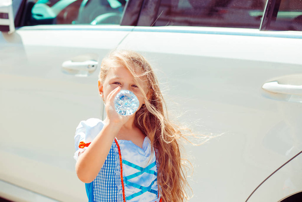Thirsty. A little girl drinking from a bottle of water, near a white car outside in summer - Fotografie, Obrázek