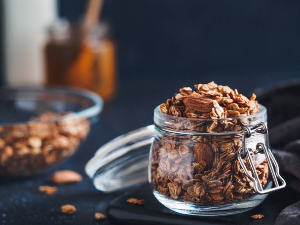 Homemade granola in glass jar on dark table. Ingredients for healthy breakfast - granola, milk and honey. Copy space for text. Low key. - Photo, image