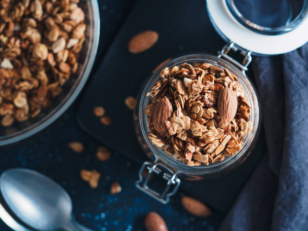 Homemade granola in glass jar on dark table. Ingredients for healthy breakfast. Copy space for text. Low key. Selective focus. Shallow DOF. Top view or flat-lay. - Foto, afbeelding