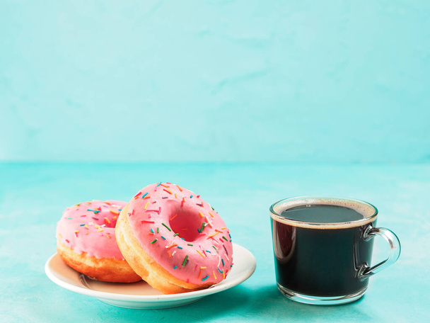 Two pink donuts and coffee on blue concrete background with copy space. Colorful donuts in plate and coffee cup with copyspace. Glazed doughnuts with sprinkles - Fotografie, Obrázek