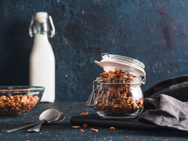 Homemade granola in glass jar on dark table. Ingredients for healthy breakfast - granola, milk and honey. Copy space for text. Low key. - Foto, afbeelding