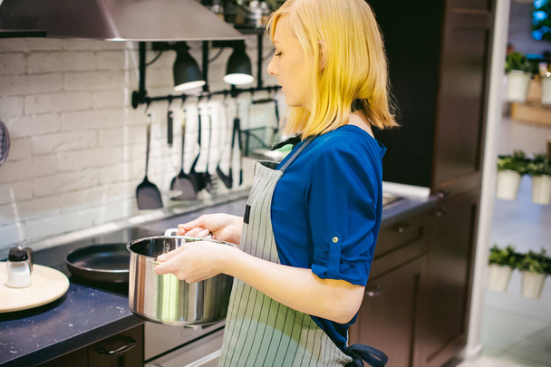 blondýnka s hrncem v kuchyni. hospodyňky v zástěře se vaří v kuchyni, dát hrnce a pánve na varnou desku - Fotografie, Obrázek