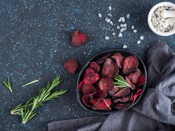 Baked beet slices with coarse sea salt scented rosemary. Vegan diet food idea and recipe. Healthy homemade beetroot chips in plate and ingredients on dark blue background. Copy space. Top view - Photo, image