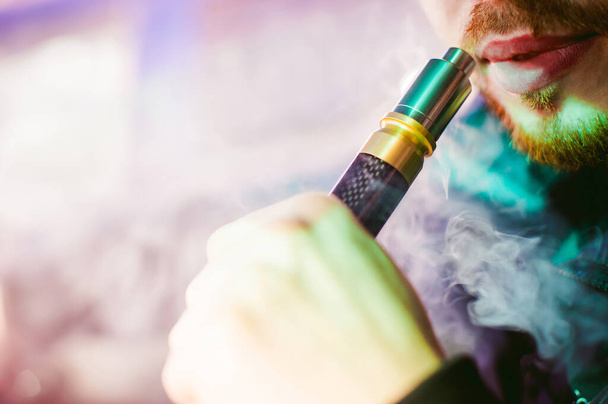 young vaper man with beard vaping mechanical mod. Guy smokes an electronic cigarette by blowing a smoke vapor. Bringing rda mouthpiece to the lips on colorful background - Foto, afbeelding