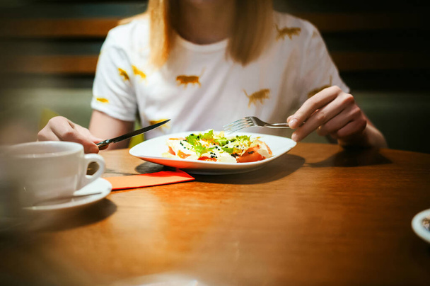 Νεαρή ξανθιά γυναίκα, φορώντας άσπρο T-shirt με στάμπα, κορίτσι τρώει σαλάτα στο μεσημεριανό γεύμα, κάθεται σε ένα τραπέζι σε ένα καφενείο, φόντο τυποποιημένο ξύλο υφή φωτίστηκε με θερμό φως - Φωτογραφία, εικόνα
