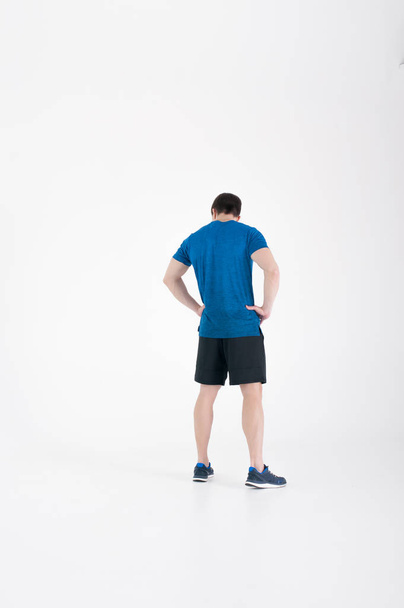 Young muscular handsome athletic man dressed in blue sport shirt, shorts and sneakers standing on white background in studio, Holding hands at waist - Foto, afbeelding