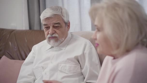 Close-up portrait of old Caucasian grey-haired man talking with wife at home. Married retirees resting together indoors in the evening. Bonding, support, love. - Video