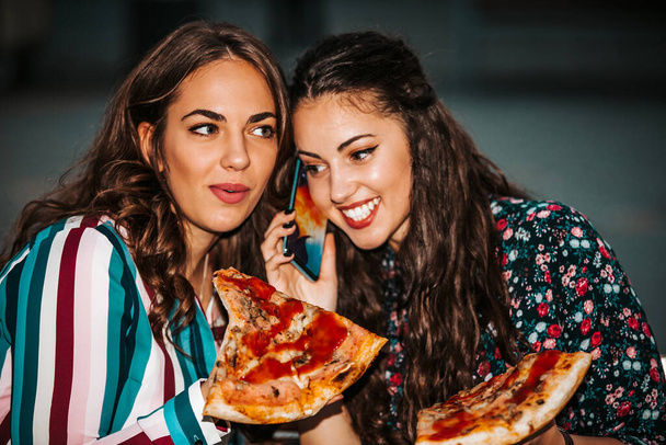 Two beautiful girls talking and eating pizza outdoors. - 写真・画像