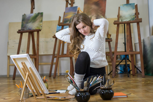 Artystka maluje i dotyka włosów siedząc na podłodze w pracowni artystycznej. - Zdjęcie, obraz