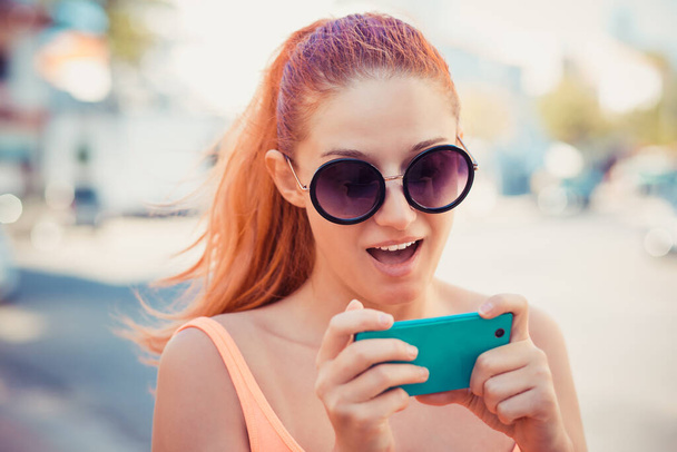 Женщина пишет смс. Крупный план молодой счастливой улыбаясь весело красивая удивленная девушка смотрит на мобильный телефон чтение отправки смс на городской пейзаж
 - Фото, изображение