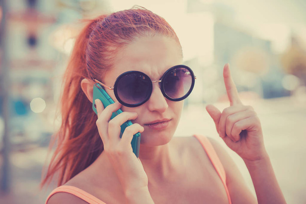 Attention, listen to me. Close up portrait of young unhappy frustrated woman wagging finger talking at phone on city scape background. - Foto, Imagen