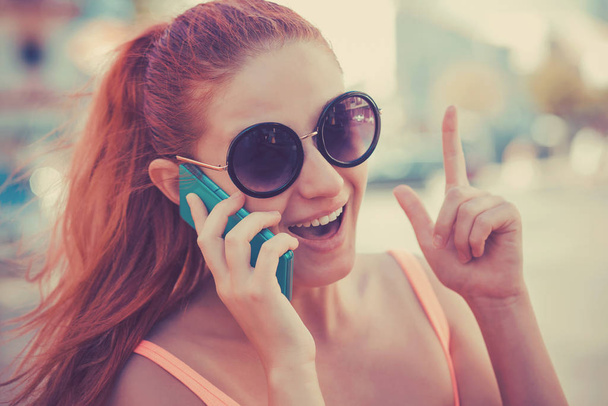 SMS. Closeup portrait happy pleased young girl having idea pointing up finger gesture talking at phone receiving good news photos message toothy smile emotion on face on cityscape - Foto, imagen