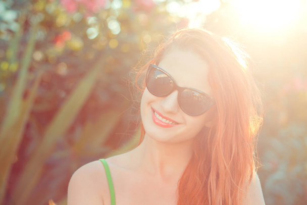 woman with sunglasses smiling on a sunny day, sunflare on right on a green bush with pink flowers on background - Foto, afbeelding