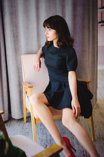 Young attractive woman with long legs in black elegant dress, sits in chair near window in interior of room. - 写真・画像