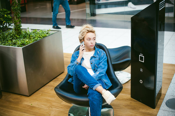 young beautiful woman in jeans clothes in business space of shopping center. portrait of a girl with freckles on her face, stylish girl sits in an armchair with a cell phone in wi-fi zone - Foto, imagen