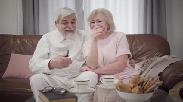 Portrait of positive mature Caucasian couple in love watching TV in the evening. Happy senior husband and wife resting together indoors. Leisure, happiness, lifestyle, eternal love concept. - Metraje, vídeo
