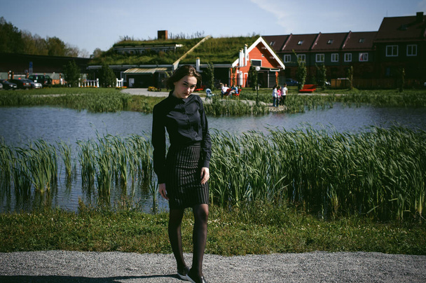 street portrait of a student girl dressed fashionably in dark clothes, against a pond background in the autumn sunny day - Foto, Imagen
