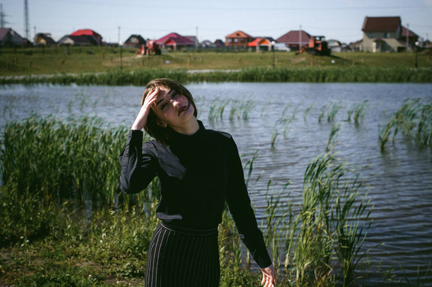 street portrait of a student girl dressed fashionably in dark clothes, against a pond background in the autumn sunny day - Photo, Image