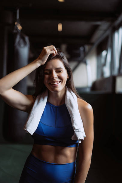 Sonriente chica en forma sosteniendo la toalla y descansando en el gimnasio
. - Foto, Imagen