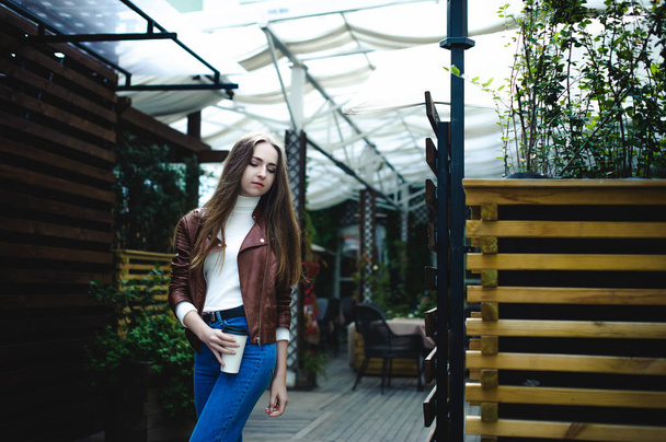 street portrait of a female student in fashionable clothes, blue jeans and a brown jacket, with a coffee on takeaway - Foto, Bild