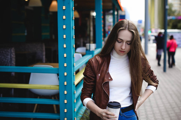 street portrait of a female student in fashionable clothes, blue jeans and a brown jacket, with a coffee on takeaway - Fotó, kép