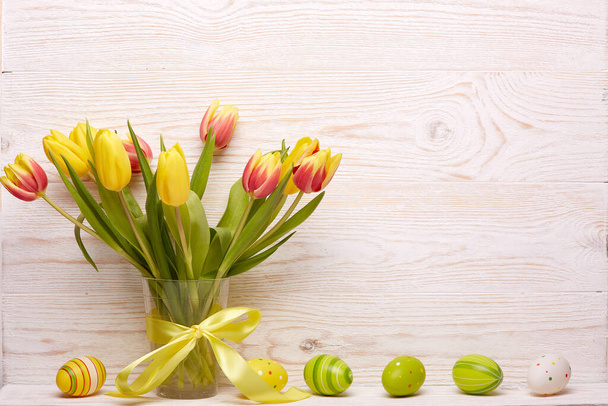 Кольорові великодні яйця, весняні тюльпани квіти у вазі та стрічки на дерев'яному тлі. Вітальна листівка
. - Фото, зображення