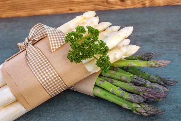 fresh green and white asparagus bunched with parsley on gray slate plate in a kitchen - Fotó, kép