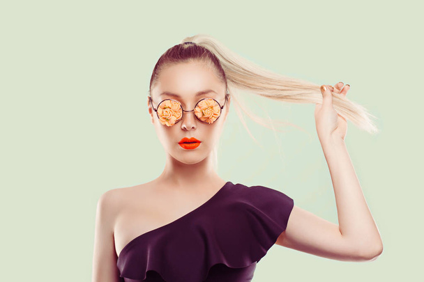 Bold stylish beautiful and trendy. Girl woman with flourish floral flower glasses combined with coral pastel lipstick, black one shoulder dress pulling her ponytail hair on light green background wall - Foto, imagen