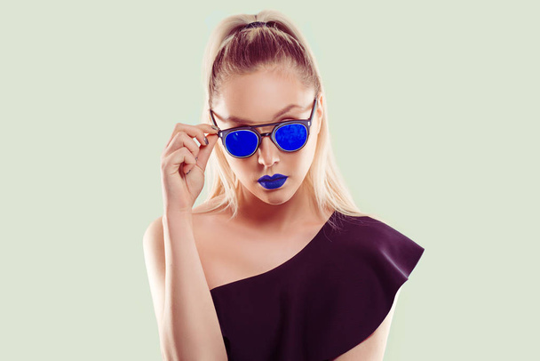 Fashioned in blue. Beautiful woman holding blue sunglasses in combination with her blue lipstick isolated light green background - Photo, image