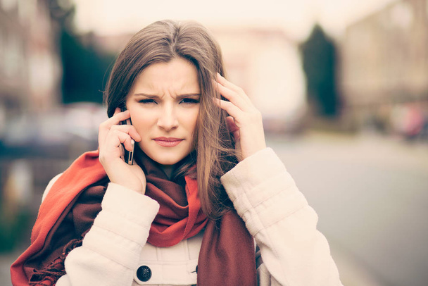 Phone talk. Portrait frustrated beautiful woman speaking on the mobile phone in a city street unfocused background having headache head pain problem stress depression hand on ear looking at you camera - Фото, зображення