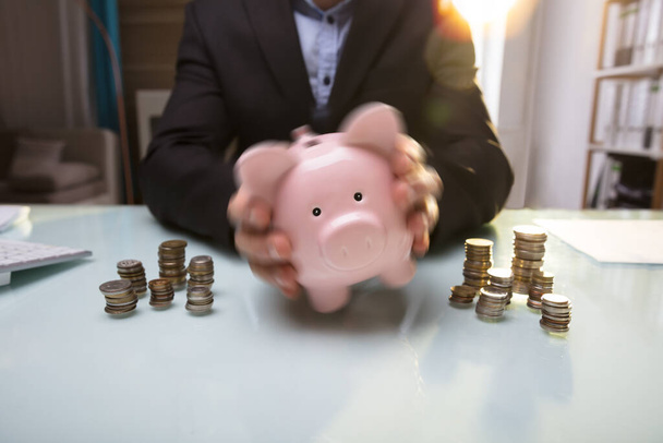Close-up Of A Businessperson's Hand Shaking Piggy Bank Near Stacked Coins - Fotoğraf, Görsel