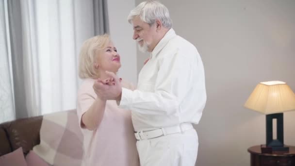 Middle shot of smiling mature Caucasian husband and wife dancing at home. Happy senior couple in love spending Saint Valentines Day indoors. Joy, happiness, romance, eternal love. - Materiaali, video