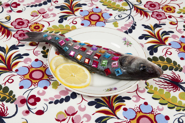 A bass fish covered with colorful precious stones inside a flower plate hidden on a pop floral patterned fabric background. Minimal quirky color still life photography. - Valokuva, kuva