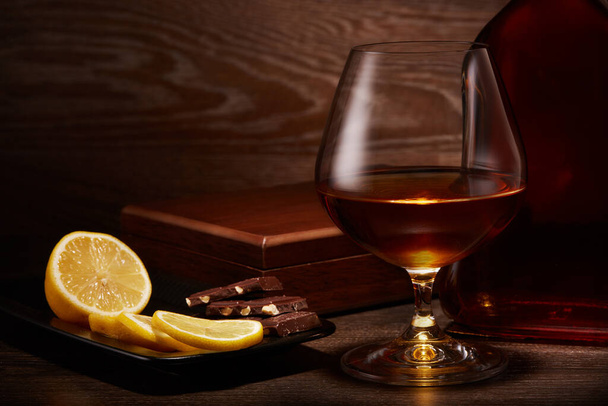 Still life with bottle of cognac, glass with cognac and plate with pieces of lemon and chocolate standing on a table on a dark wooden background. - Foto, afbeelding