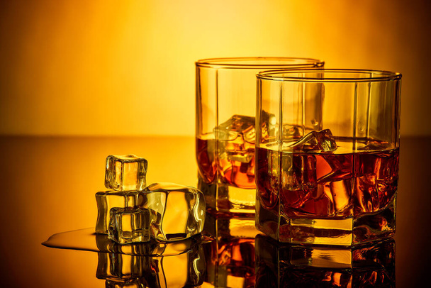 Two glasses of scotch whiskey with ice cubes standing on a reflective surface over yellow background. - Photo, image