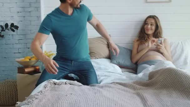 Pretty pregnant woman texting smartphone. Joyful pregnant couple lying in bed - Video, Çekim