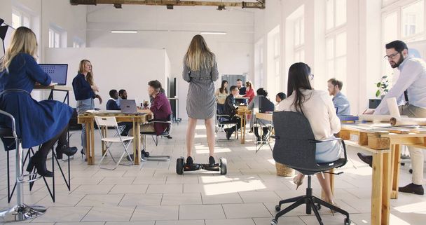 Cámara sigue confiado líder femenino utilizando scooter eléctrico para moverse por la oficina loft moderno. Lugar de trabajo saludable
. - Foto, imagen