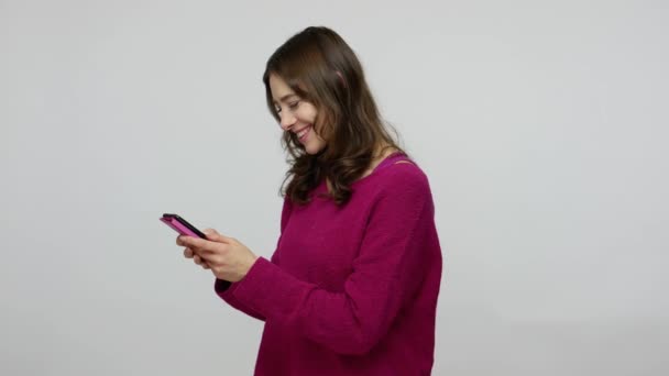 Happy charming brunette woman in pullover using mobile phone, typing message on social network and laughing - Materiał filmowy, wideo