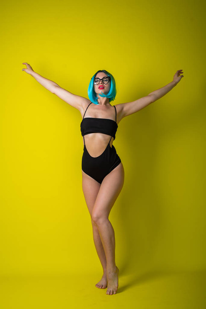 Beautiful woman in a black bikini in a colored wig and sunglasses on a yellow background. Girl posing in the studio. - Foto, afbeelding