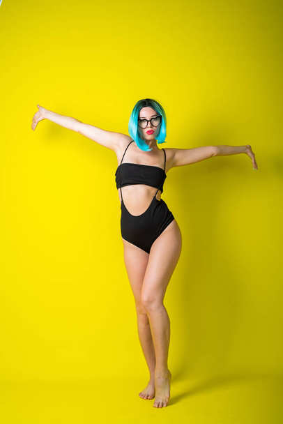Beautiful woman in a black bikini in a colored wig and sunglasses on a yellow background. Girl posing in the studio. - Photo, image