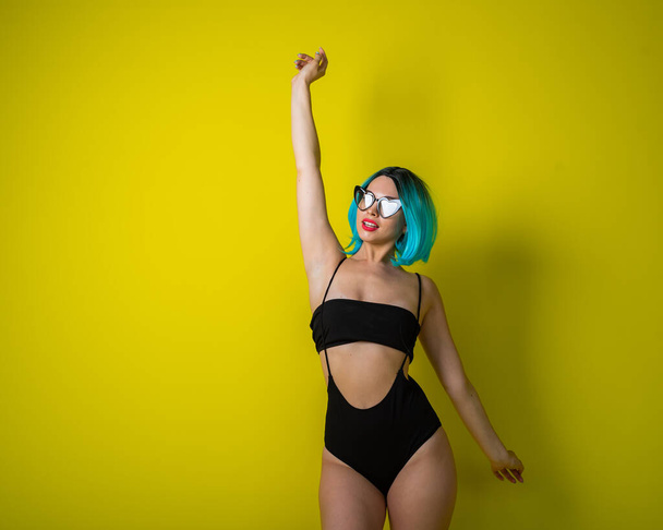 Beautiful woman in a black bikini in a colored wig and sunglasses on a yellow background. Girl posing in the studio. - Фото, зображення